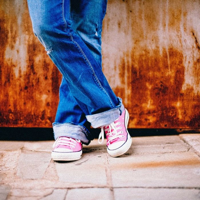 bottom half of a person in blue denim jeans and pink and white nike sneakers standing against a wall with their legs crosssed.