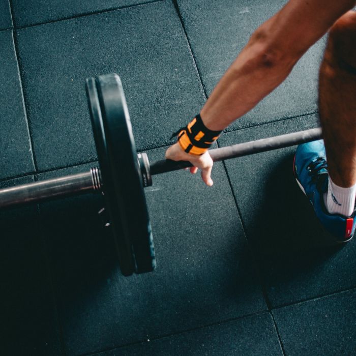 A photograph of a person in a gym holding a barbell, his arm and leg visible in the frame.