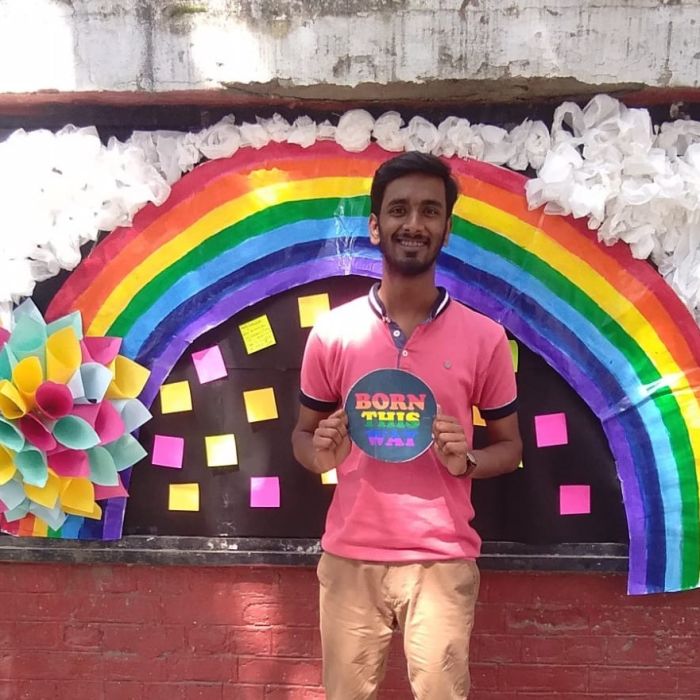 photo of Rajeev in a pink tee-shirt and light brown pants and holding a sign saying 'Born this way'. They are standing in front of a pride rainbow pasted on a wall. There are colourful post-its on a black patch on the wall.