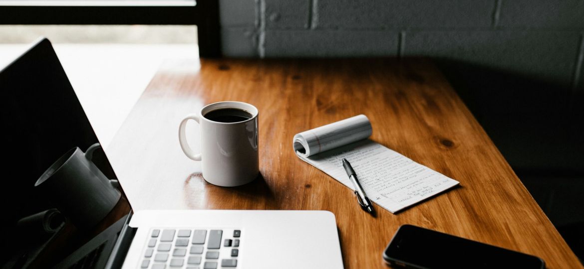 A MacBook Pro, a white ceramic mug, an open notepad with a pen, and a black smartphone on a brown wooden table.