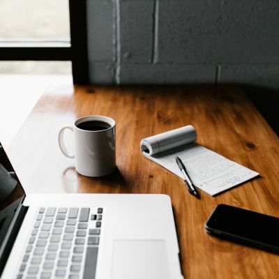 A MacBook Pro, a white ceramic mug, an open notepad with a pen, and a black smartphone on a brown wooden table.