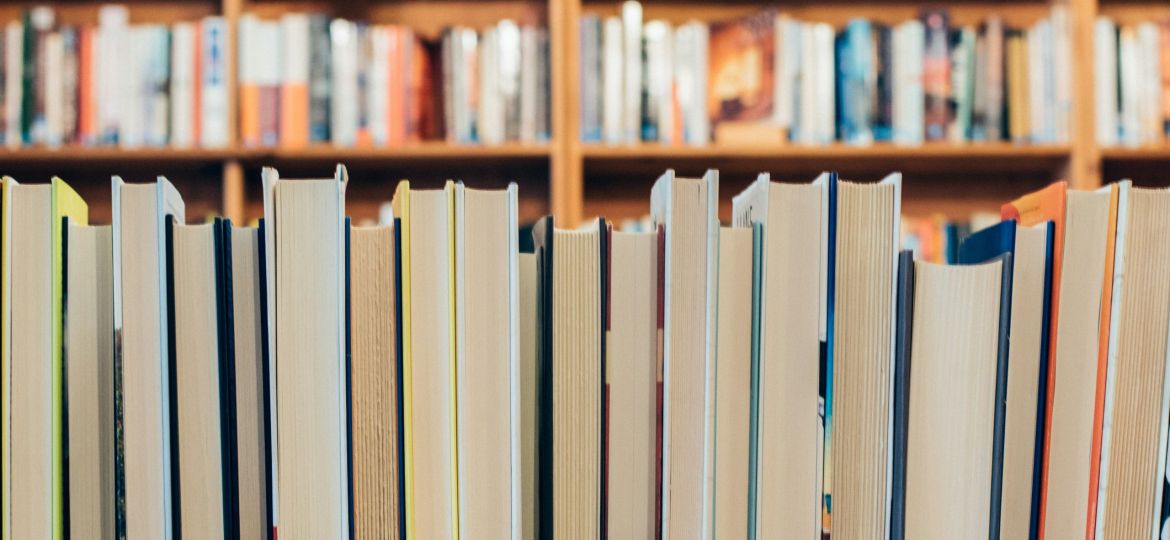 A row of assorted books in focus with the spines facing away from the camera. In the background, there are many shelves filled with books.