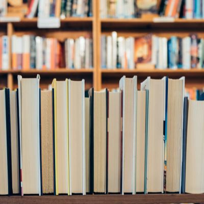 A row of assorted books in focus with the spines facing away from the camera. In the background, there are many shelves filled with books.