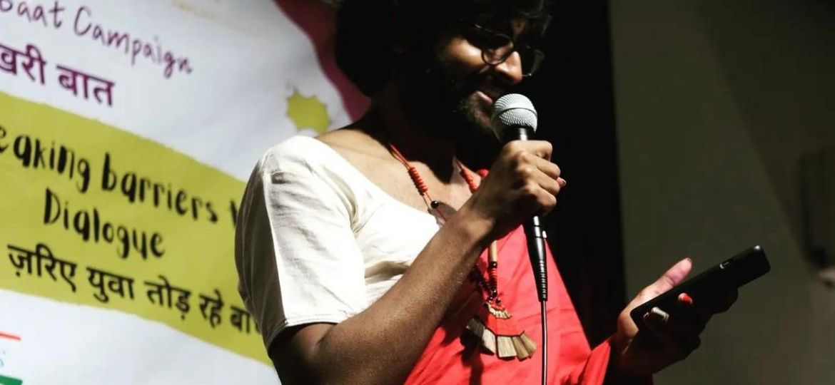 A photo of Chand dressed in a red sari with a white blouse, holding a mike on a stage and reading out from their phone.