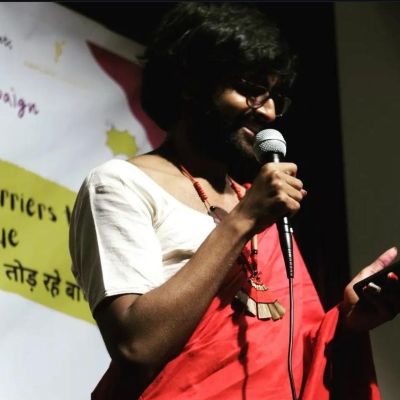 A photo of Chand dressed in a red sari with a white blouse, holding a mike on a stage and reading out from their phone.