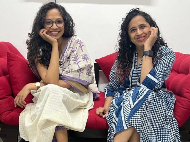 Image of Vani and Ramya seated on a red sofa. Vani is in a white kurta and a dupatta with purple flowers. Ramya is in a blue and white patterned dress. Both have their hands on their chins and are smiling.